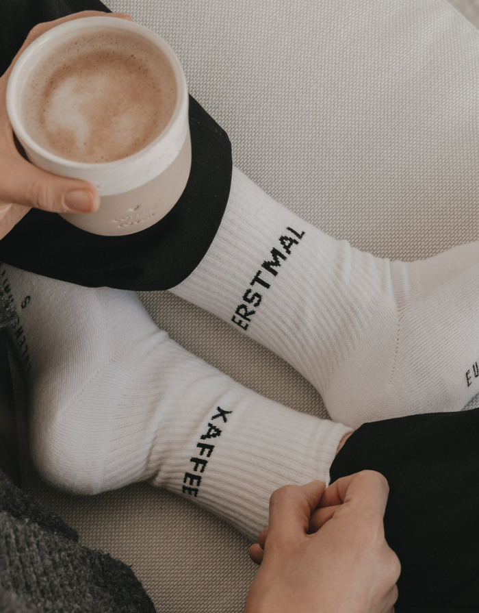 Weiße Socken Erstmal Kaffee von Eulenschnitt mit schwarzem Schriftzug an der Ferse und schwarzen Details