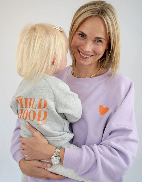 Liliput Motherhood Oversize Sweatshirt für Damen mit kleinem orangefarbenem Herz auf der Vorderseite und großem Motherhood-Schriftzug auf dem Rücken, getragen.