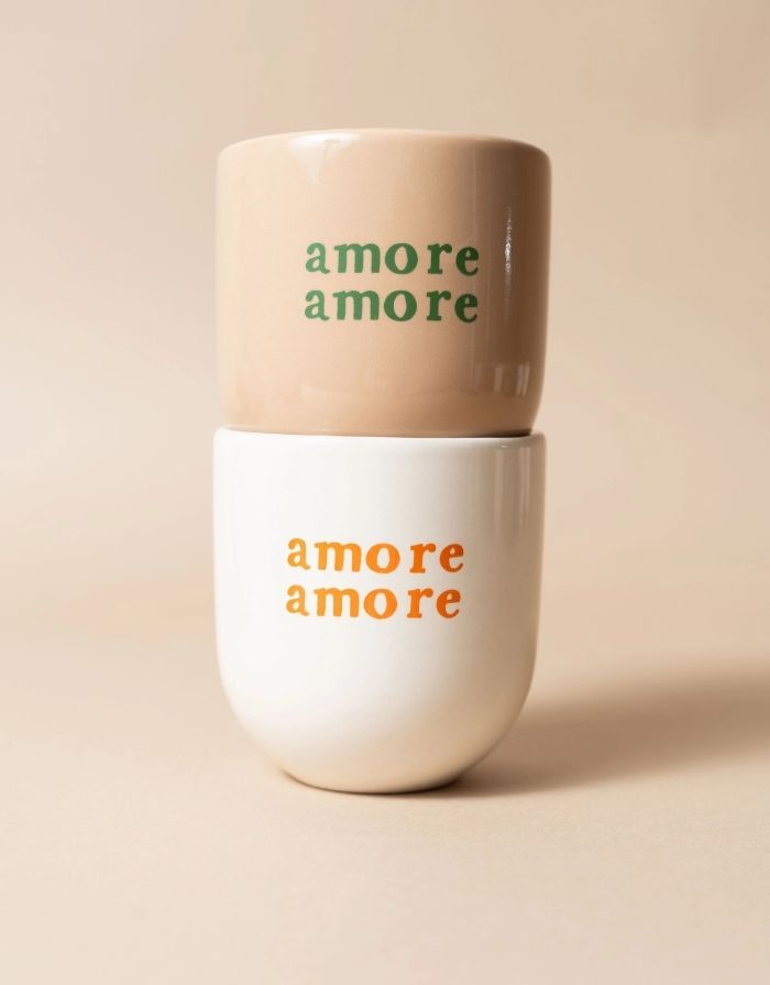 Becher von Sisi House of Style in Beige mit grünem amore amore Schriftzug auf einem weißen Becher mit orangenem amore amore Schriftzug 