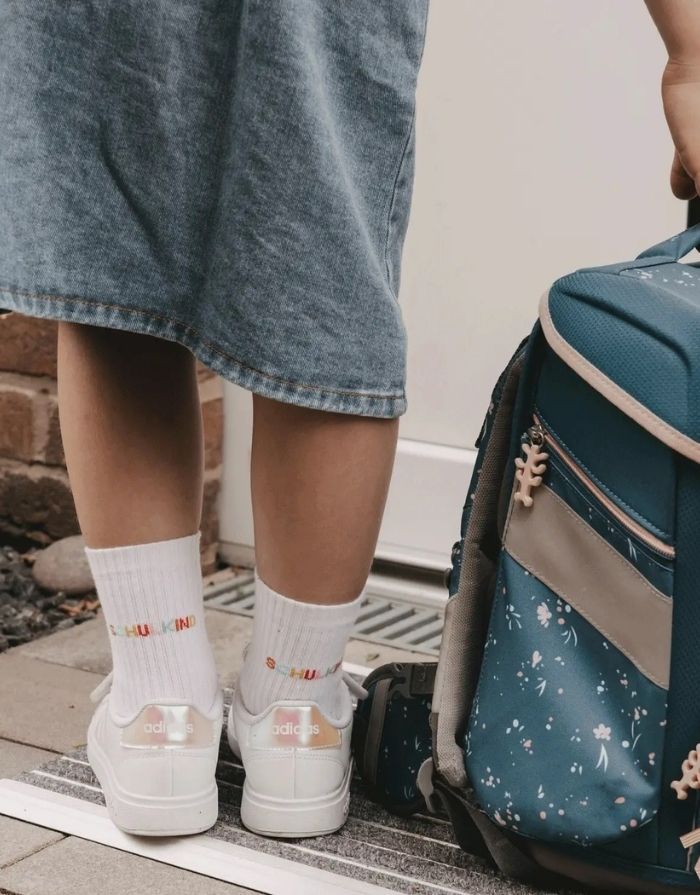 Weiße Eulenschnitt Socken „Schulkind“ mit buntem Schriftzug auf der Ferse und weichen Bündchen für Kinder an einem Kind von hinten mit Schulranzen