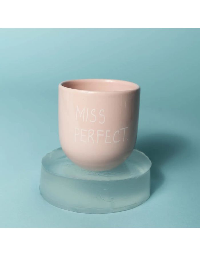 Becher Miss Perfect von SISI House of Style – zartrosa Keramik mit weißem Schriftzug, auf Glassockel vor hellblauem Hintergrund
