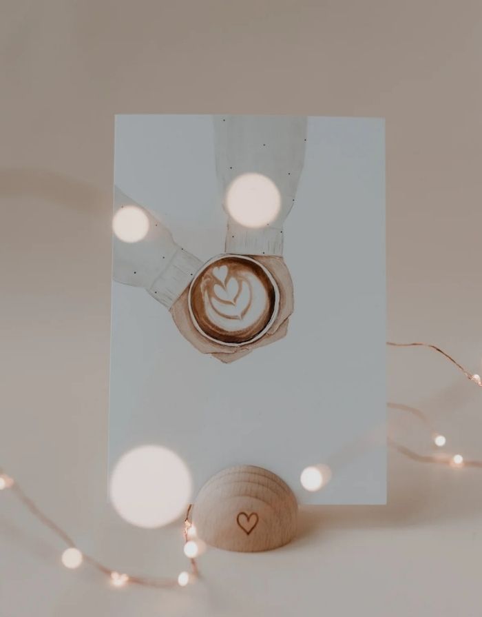 Postkarte in Weiß von Eulenschnitt in Kartenständer mit Händen die einen Kaffee halten